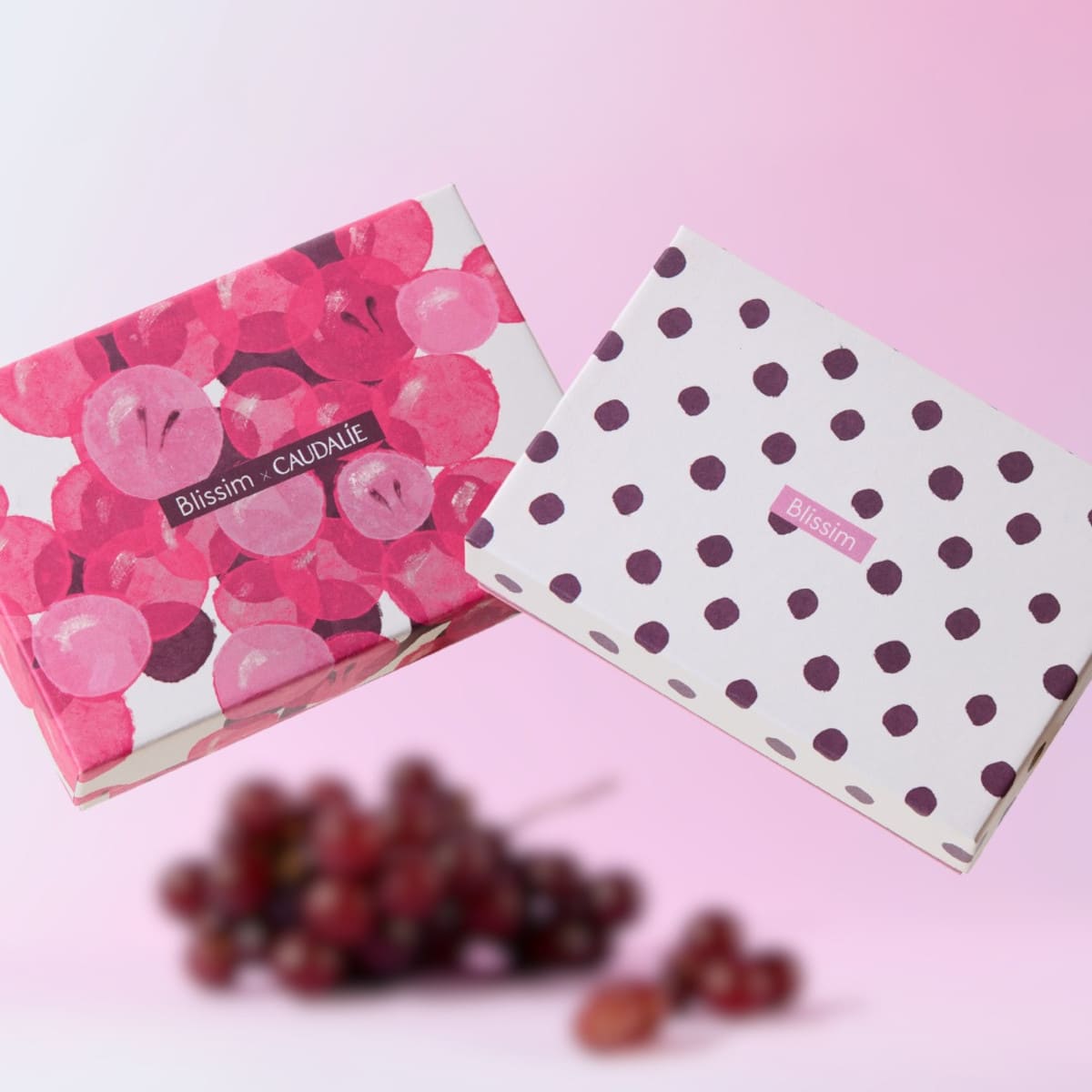 Deux box Blissim sur fond rose, avec une édition Caudalie à motifs de raisins et une box blanche à pois