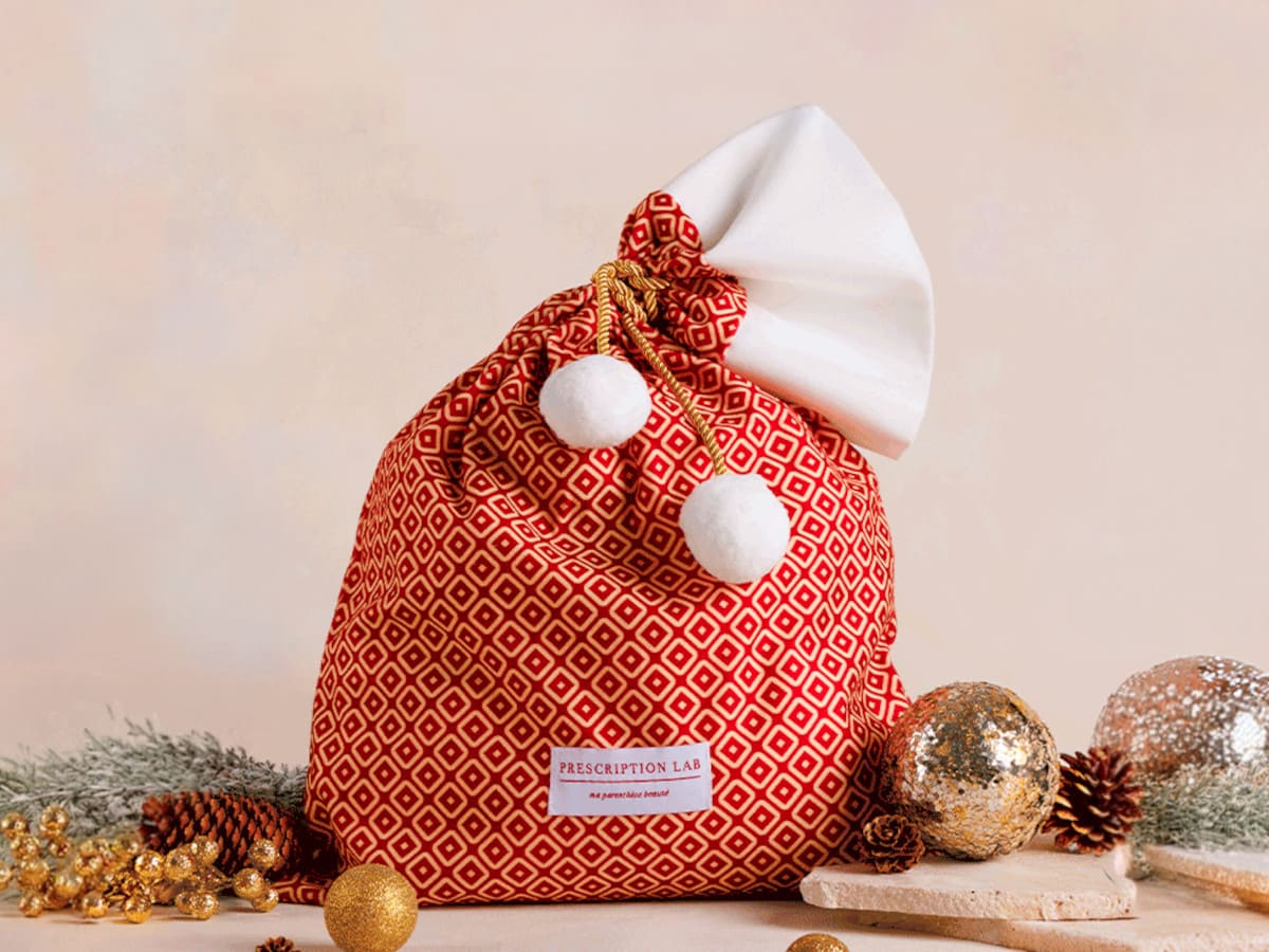 Hotte de Noël Prescription Lab en tissu rouge à motifs géométriques, avec cordon doré et pompons blancs.