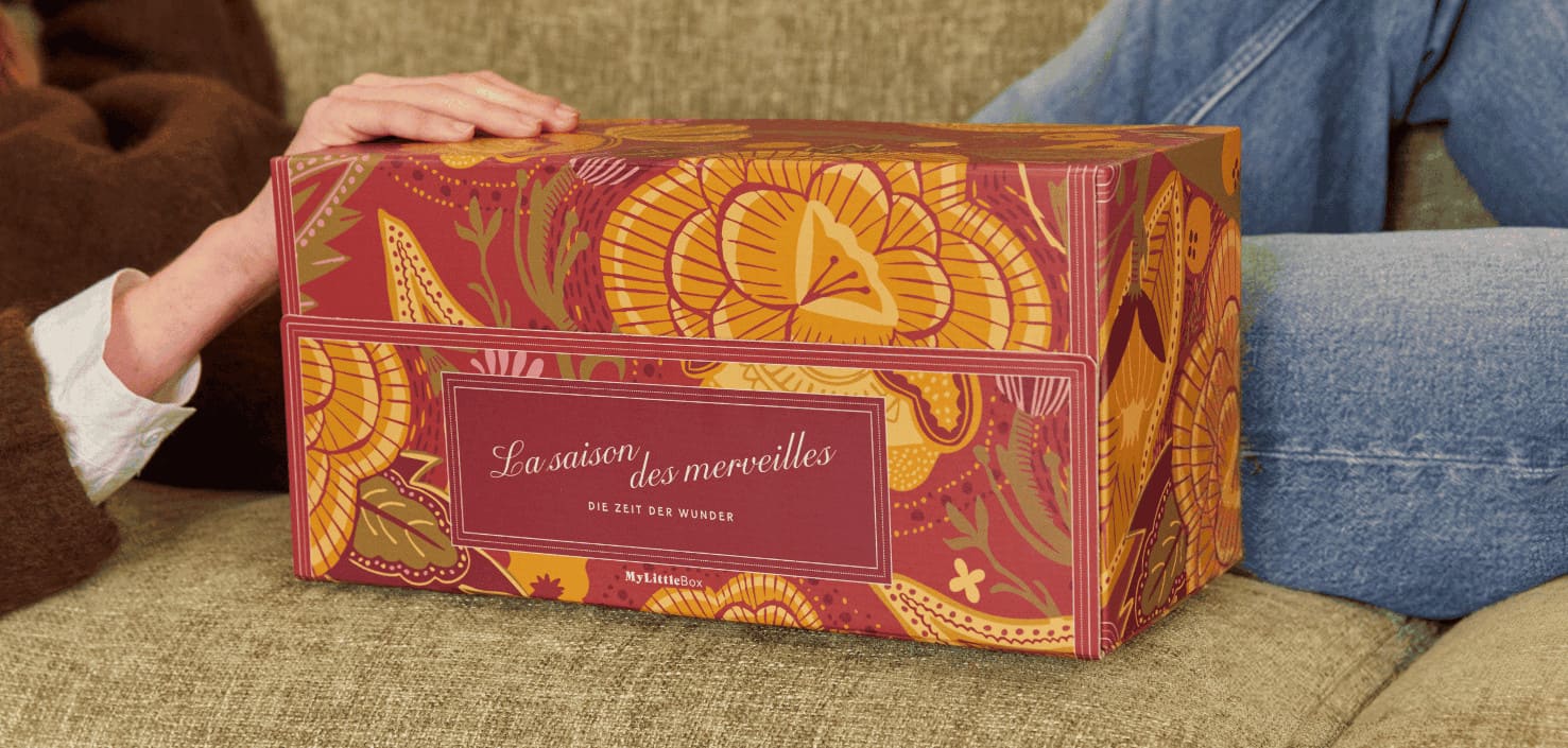 Une femme assise sur un canapé présente le coffret Les Merveilles de Noël de My Little Box, aux illustrations florales colorées