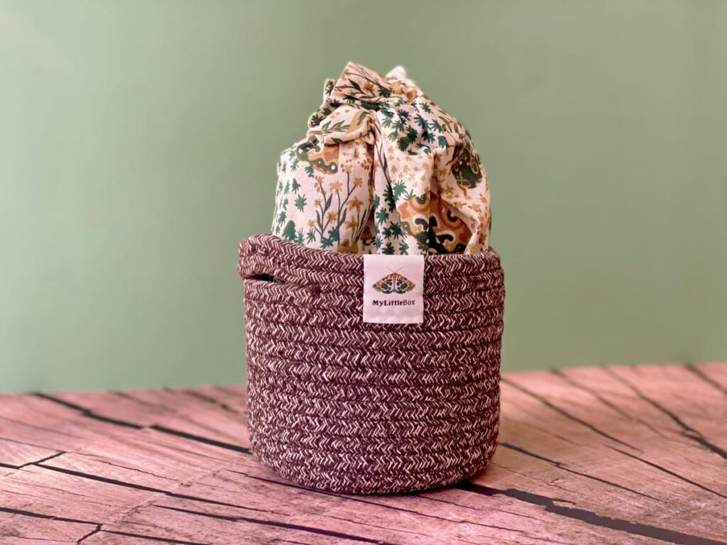 Panière tressée de l'édition Murmures des Bois avec son pochon en tissu aux motifs végétaux, photographiée sur une table en bois.